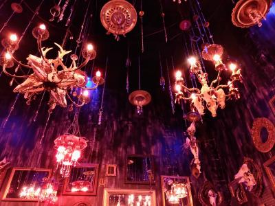 An installation with chandeliers hanging from the ceiling of a room. The room is dim lit, and there are mirrors hanging on its walls to heighten the effect of the hanging chandeliers.